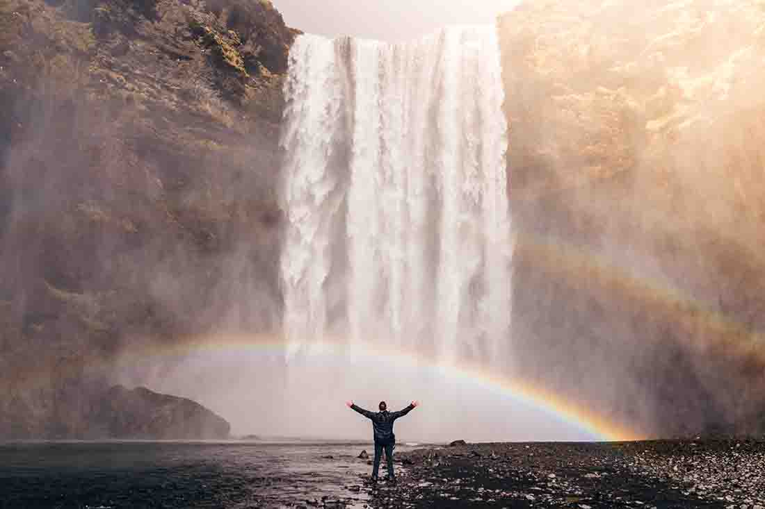 Man standing near a giant waterfall with a rainbow at the bottom left