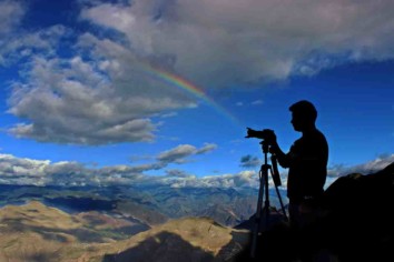 a photographer with a camera on a mountain top with clouds and a rainbow in the background